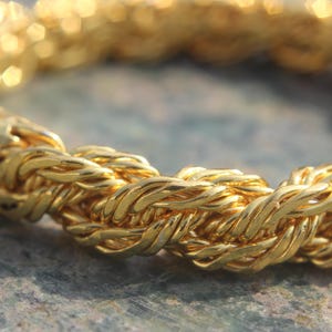 Könnte beinhalten: Nahaufnahme eines goldfarbenen Armbands mit einem seilartigen Design. Das Armband besteht aus ineinander verschlungenen Strängen, wodurch ein strukturiertes Aussehen entsteht. Der Schmuck befindet sich vor einem verschwommenen Hintergrund.