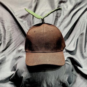 May include: A brown baseball cap with a green sprout design. The cap has a curved brim and a fabric texture. The sprout is made of green fabric and is attached to the top of the cap.