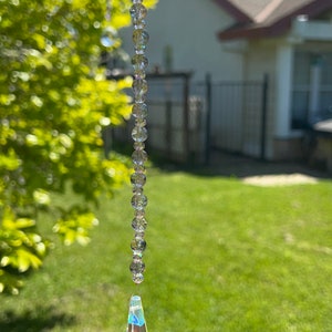 May include: A crystal suncatcher with a teardrop-shaped prism hanging from a chain of clear beads. The suncatcher is hanging from a tree branch in a backyard setting.