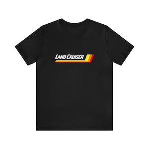 May include: Black t-shirt featuring the text "LAND CRUISER" in white, with a horizontal stripe design in yellow, red, and orange. The shirt is a short-sleeved crew neck.