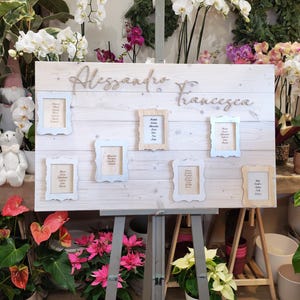May include: A wooden sign with the names "Alessandro & Francesca" in cursive script. The sign has several small framed cards with names, and is displayed on a gray easel. The background includes various plants and flowers.