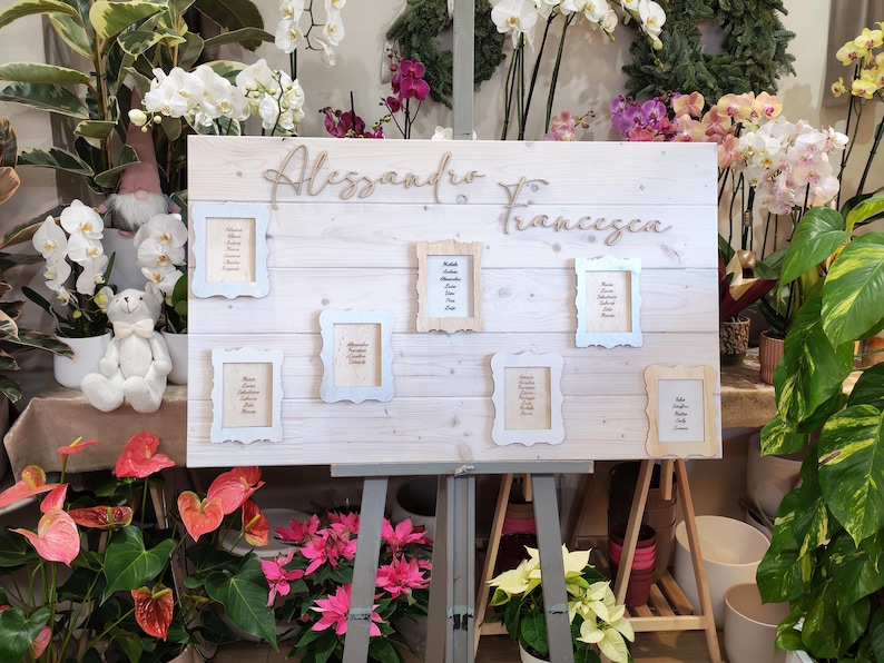 May include: A wooden sign with the names "Alessandro & Francesca" in cursive script. The sign has several small framed cards with names, and is displayed on a gray easel. The background includes various plants and flowers.