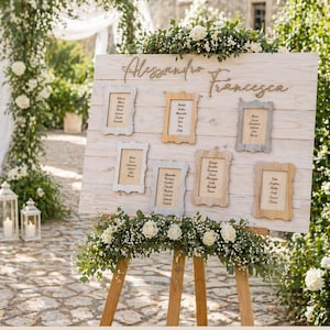 May include: A wooden wedding seating chart with the names "Alessandro Francesca" in script. The chart has six framed cards with guest names, and is decorated with white flowers and greenery. The sign is on a wooden easel.