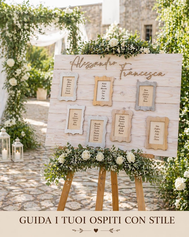 May include: A wooden wedding seating chart with the names "Alessandro Francesca" in script. The chart has six framed cards with guest names, and is decorated with white flowers and greenery. The sign is on a wooden easel.