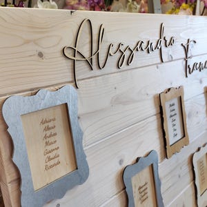 May include: Wooden wedding seating chart with the names "Alessandro" and "Francesca" in cursive script. Several small framed cards with guest names are attached to the light-colored wooden board. The frames are in silver and brown.