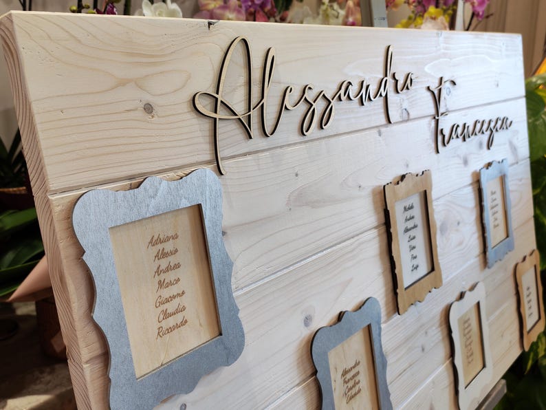 May include: Wooden wedding seating chart with the names "Alessandro" and "Francesca" in cursive script. Several small framed cards with guest names are attached to the light-colored wooden board. The frames are in silver and brown.