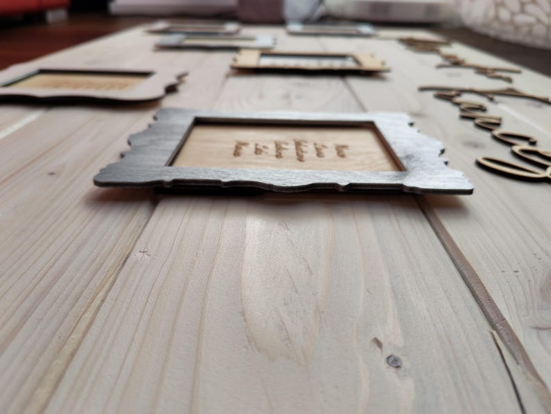 May include: Wooden framed signs with engraved text, arranged on a light wood surface. The frames have a dark gray border and a natural wood interior. The word "coffee" is engraved on one sign.