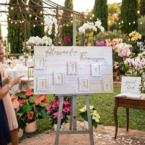 May include: A rustic wooden wedding seating chart on an easel, with the names "Alessandro" and "Francesca" in cursive. The chart features framed table assignments and is decorated with white orchids. A guest book table is nearby.