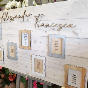 May include: A wooden wedding seating chart with the names "Alessandro" and "Francesca" in cursive script. Several small framed cards with guest names are attached to the light-colored wooden board. The board is propped up on a metal stand.