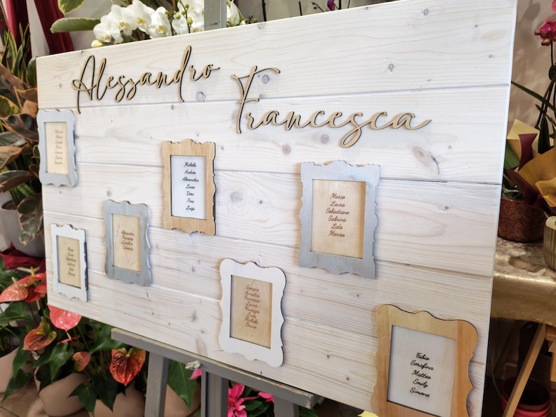 May include: A wooden wedding seating chart with the names "Alessandro" and "Francesca" in cursive script. Several small framed cards with guest names are attached to the light-colored wooden board. The board is propped up on a metal stand.