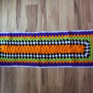 May include: A rectangular, handmade crochet table runner. The runner features a vibrant color scheme with a purple border, green and white accents, and an orange center. The edges are finished with a white border.