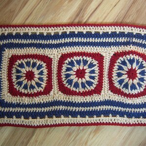 May include: A rectangular, handmade crochet piece with a repeating pattern. The design features red, blue, and cream-colored squares and borders. The central squares have a starburst design. The piece is likely a table runner or decorative mat.