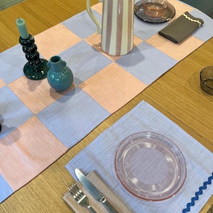 Könnte beinhalten: Eine Tischdekoration mit einem rosa-blauen karierten Tischläufer. Eine gestreifte Kanne mit gelbem Griff steht auf dem Läufer, zusammen mit einem Kerzenhalter, einer kleinen Vase und einem Teller. Ein Gedeck mit Besteck und einer Serviette ist ebenfalls zu sehen.