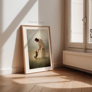 May include: A framed print titled "Paw of Love" shows a figure in a white robe holding the paw of a small dog. The artwork is set in a field with soft, diffused light, creating a serene atmosphere. The frame is a light wood colour.