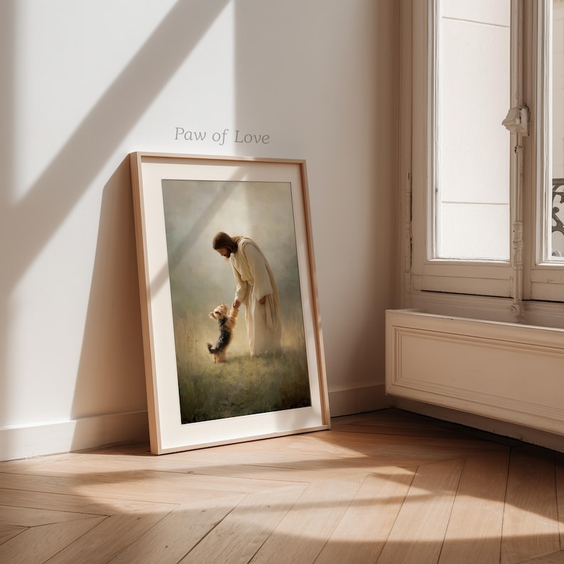 May include: A framed print titled "Paw of Love" shows a figure in a white robe holding the paw of a small dog. The artwork is set in a field with soft, diffused light, creating a serene atmosphere. The frame is a light wood colour.