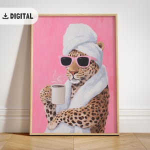 Op de afbeelding: Digitale print van een luipaard met een witte handdoek en roze zonnebril, die een dampende koffiemok vasthoudt. De afbeelding is omlijst in een licht houten frame op een roze achtergrond. De afbeelding heeft een "DIGITAL" download-pictogram.