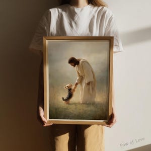 May include: A framed print shows a figure in white robes interacting with a small dog in a grassy field. The artwork has a soft, diffused light, creating a peaceful atmosphere. The frame is made of light-coloured wood. The print is a Paw of Love design.