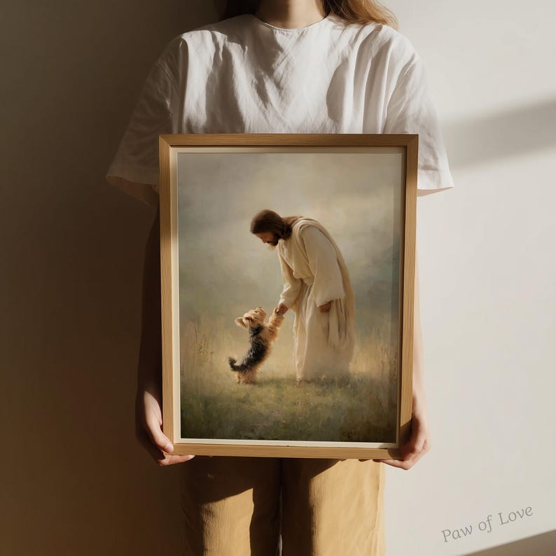 May include: A framed print shows a figure in white robes interacting with a small dog in a grassy field. The artwork has a soft, diffused light, creating a peaceful atmosphere. The frame is made of light-coloured wood. The print is a Paw of Love design.