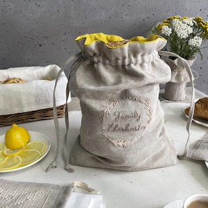 May include: A beige linen drawstring bag with a bright yellow interior lining. The front features an embroidered circular wreath design surrounding the text "Family Eberharter" in a cursive script. The bag has two long matching beige cords.