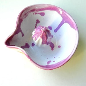 Puede incluir: Exprimidor de cítricos de cerámica hecho a mano con interior blanco y exterior rosa y morado. El exprimidor tiene un pico y un escariador en el centro. Salpicaduras de pintura rosa decoran el cuenco.