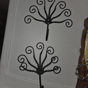 May include: Two black metal wall hooks with a decorative, stylized floral design. Each hook features a central circular element with radiating curved lines ending in swirls. The hooks are mounted on a white surface.