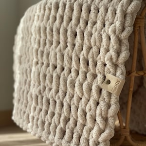 Op de afbeelding: Een chunky gebreide deken in een neutrale beige kleur, gedrapeerd over een rieten mand. De deken heeft een dikke, getextureerde weefsel en een klein leren label met de tekst "MyGile" is bevestigd. De deken ziet er zacht en gezellig uit.