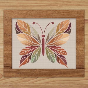 May include: A framed cross-stitch depicting a butterfly with wings resembling leaves. The wings are a mosaic of autumnal hues: orange, red, green, and brown. The frame is wooden, and the background is a neutral beige.