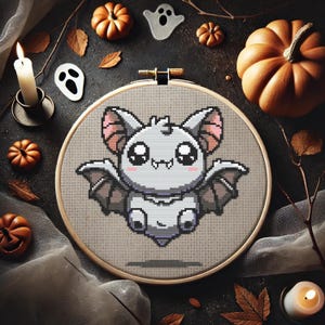May include: A cross-stitch embroidery hoop with a cartoon bat design, surrounded by Halloween decorations. The bat is grey with pink details. The scene includes pumpkins, leaves, ghosts, and a lit candle, creating a spooky theme.