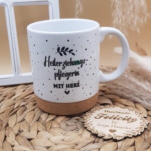 Könnte beinhalten: Weißer Keramikbecher mit Korkboden, mit schwarzem Text und kleinen schwarzen Punkten. Der Text lautet "Heilerziehungspflegerin MIT HERZ." Ein Holzschild mit "Handmade Felicity by Anja Haucke" ist ebenfalls sichtbar.
