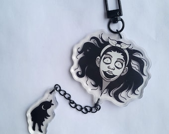 May include: A black and white acrylic keychain featuring a cartoon character with long hair and a surprised expression. The keychain has a black chain and a small black and white charm.