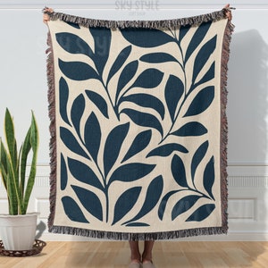 May include: A woven throw blanket with a cream background and a navy blue leaf pattern. The blanket has fringe on all sides.