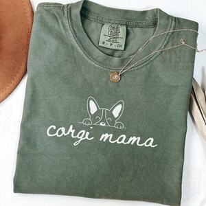 May include: A green t-shirt with a white graphic of a corgi peeking out from behind a sign that says "corgi mama".
