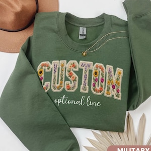May include: A military green long-sleeve sweatshirt featuring the word CUSTOM embroidered in a floral pattern with various colorful flowers. Below the word, the text optional line is printed in white cursive script. The garment has a crew neckline.