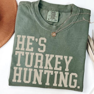 Puede incluir: Una camiseta verde con el texto "He's Turkey Hunting." impreso en beige.