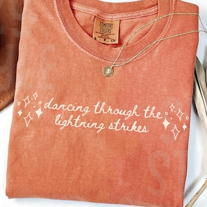 May include: A coral-colored t-shirt with the phrase "dancing through the lightning strikes" in white script. A gold necklace with a pendant rests on the shirt. A brown hat and dried plant are in the background.