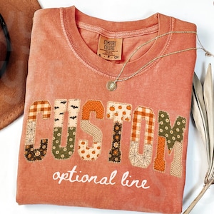 May include: A terracotta-colored t-shirt featuring the word CUSTOM in large, patchwork-style letters with various autumn-themed patterns, including pumpkins, plaid, and floral prints. Below the word, the text optional line is printed in white.