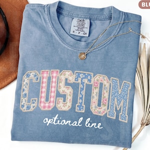 May include: Blue jean-colored t-shirt with the word "CUSTOM" in a floral and gingham design. The shirt has an optional line of text below the word "CUSTOM". A gold necklace and a brown hat are also in the image.