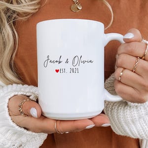 K&ouml;nnte beinhalten: Eine wei&szlig;e Keramik-Tasse mit den Namen "Jacob & Olivia" in Schreibschrift, einem roten Herz und "EST. 2021". Die Tasse hat einen gro&szlig;en Henkel. Die Tasse wird von einer Person gehalten, die Ringe und einen wei&szlig;en Pullover tr&auml;gt. Ein Geschenk f&uuml;r ein Paar.