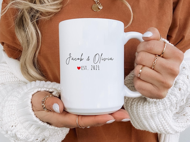 K&ouml;nnte beinhalten: Eine wei&szlig;e Keramik-Tasse mit den Namen "Jacob & Olivia" in Schreibschrift, einem roten Herz und "EST. 2021". Die Tasse hat einen gro&szlig;en Henkel. Die Tasse wird von einer Person gehalten, die Ringe und einen wei&szlig;en Pullover tr&auml;gt. Ein Geschenk f&uuml;r ein Paar.