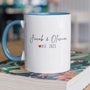 K&ouml;nnte beinhalten: Wei&szlig;er Keramikbecher mit blauem Henkel und Innenraum. Der Becher tr&auml;gt die Namen "Jacob & Olivia" in Schreibschrift, ein rotes Herz und "EST. 2021". Der Becher steht auf einem Buch mit floralem Design.
