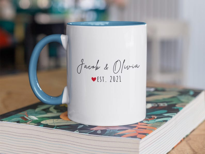 K&ouml;nnte beinhalten: Wei&szlig;er Keramikbecher mit blauem Henkel und Innenraum. Der Becher tr&auml;gt die Namen "Jacob & Olivia" in Schreibschrift, ein rotes Herz und "EST. 2021". Der Becher steht auf einem Buch mit floralem Design.
