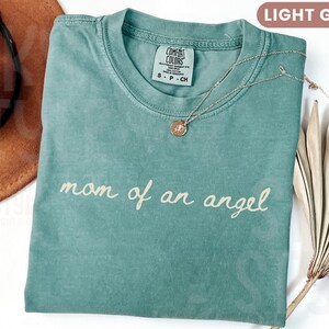 K&ouml;nnte beinhalten: Hellgr&uuml;nes T-Shirt mit dem kursiven Schriftzug "mom of an angel". Eine goldfarbene Halskette mit Anh&auml;nger liegt auf dem Shirt. Ein brauner Hut und eine getrocknete Pflanze befinden sich im Hintergrund. Das Shirt hat ein Etikett mit der Aufschrift "Comfort Colors".