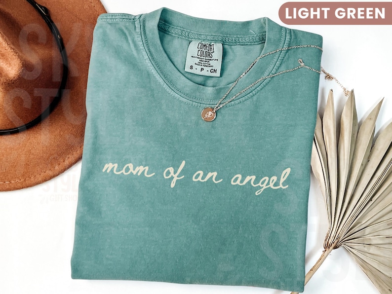 K&ouml;nnte beinhalten: Hellgr&uuml;nes T-Shirt mit dem kursiven Schriftzug "mom of an angel". Eine goldfarbene Halskette mit Anh&auml;nger liegt auf dem Shirt. Ein brauner Hut und eine getrocknete Pflanze befinden sich im Hintergrund. Das Shirt hat ein Etikett mit der Aufschrift "Comfort Colors".