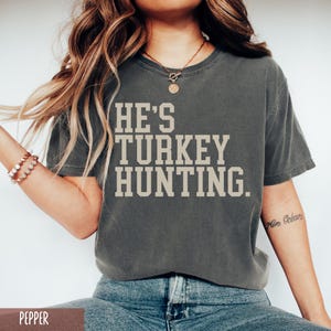 Puede incluir: Una camiseta gris con el texto "HE'S TURKEY HUNTING." impreso en blanco.