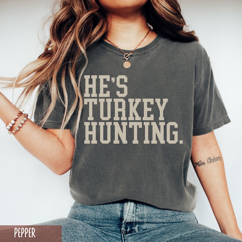 Puede incluir: Una camiseta gris con el texto "HE'S TURKEY HUNTING." impreso en blanco.