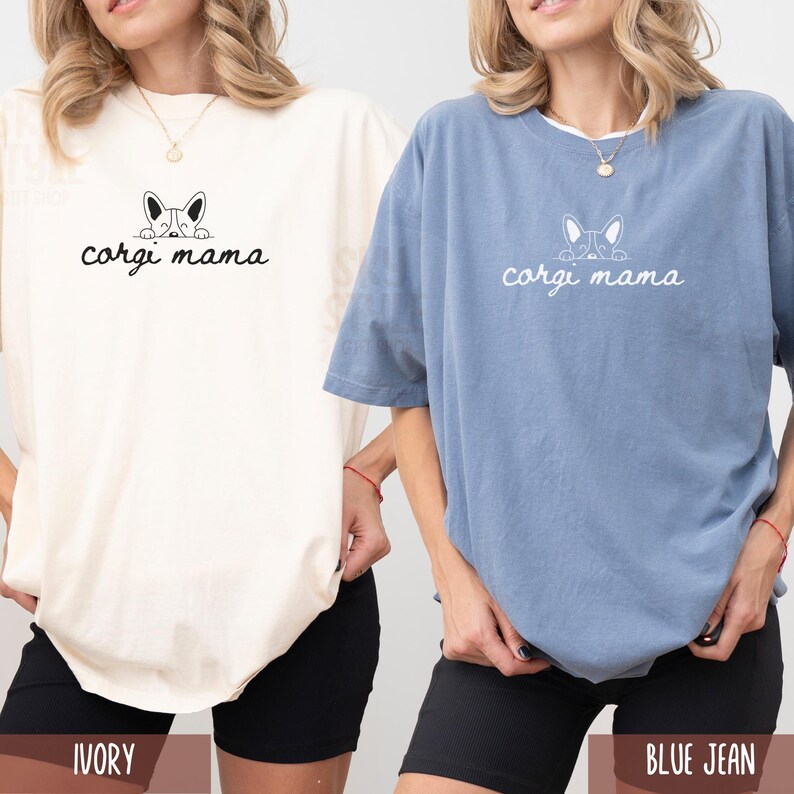 May include: Two women wearing t-shirts with the text "corgi mama" and a cartoon corgi peeking out from behind the text. The t-shirt on the left is ivory and the t-shirt on the right is blue jean.