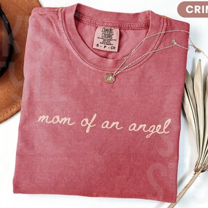 K&ouml;nnte beinhalten: Karmesinrotes T-Shirt mit dem Schriftzug "mom of an angel" in Schreibschrift. Eine goldene Anh&auml;ngerkette und ein brauner Hut sind ebenfalls zu sehen. Das Shirt ist gefaltet, und ein getrocknetes Palmblatt ist im Hintergrund.
