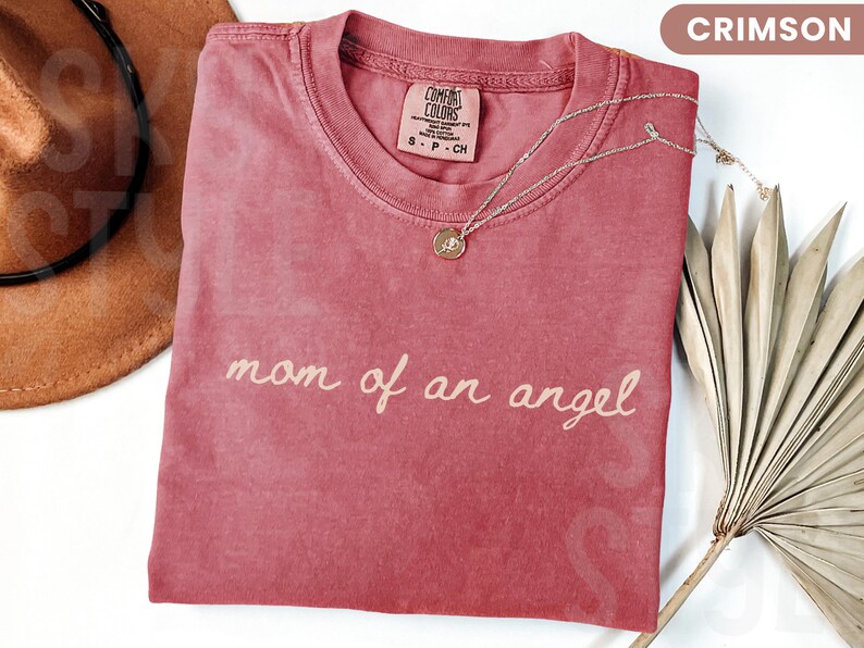 K&ouml;nnte beinhalten: Karmesinrotes T-Shirt mit dem Schriftzug "mom of an angel" in Schreibschrift. Eine goldene Anh&auml;ngerkette und ein brauner Hut sind ebenfalls zu sehen. Das Shirt ist gefaltet, und ein getrocknetes Palmblatt ist im Hintergrund.
