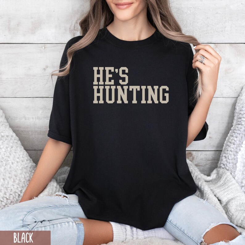 May include: Black t-shirt with the text "HE'S HUNTING" in a beige color.