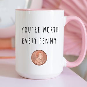 May include: White ceramic mug with a pink handle and interior. The mug features the text "YOU'RE WORTH EVERY PENNY" above a penny illustration. The mug is on a pink surface.